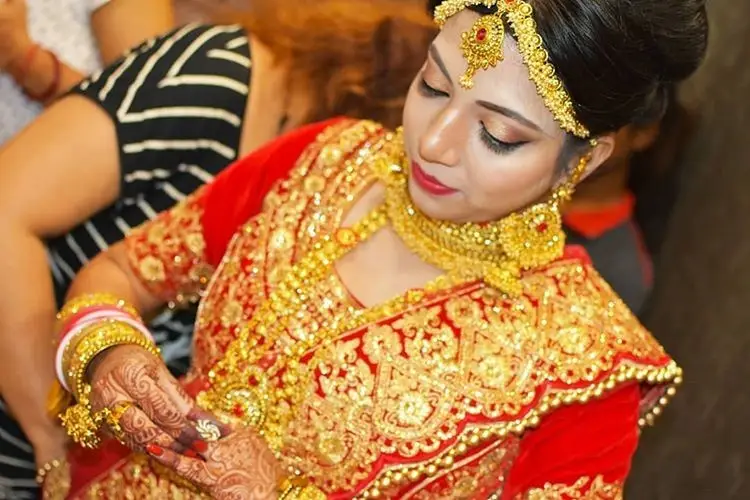 Bride in a red and gold traditional outfit with heavy gold jewelry and detailed mehndi, looking down while adjusting her hands.
