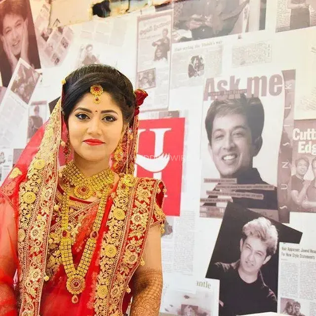 Bride in a red and gold traditional outfit with ornate jewelry and a veil, standing in front of a wall decorated with posters and photos.